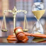 Legal office of lawyers, justice and law concept : Judge's gavel or hammer and base used by a judge person on a desk in a courtroom with blurred weight scale of justice, bookshelf, hourglass behind.