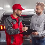 Professional mechanic making notes in checklist document while discussing car repair with male client in modern workshop