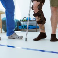 Man after car accident in an orthosis and on crutches learning to walk in the clinic, helpful therapist near him Man after car accident in an orthosis and on crutches learning to walk in the clinic, helpful therapist near him